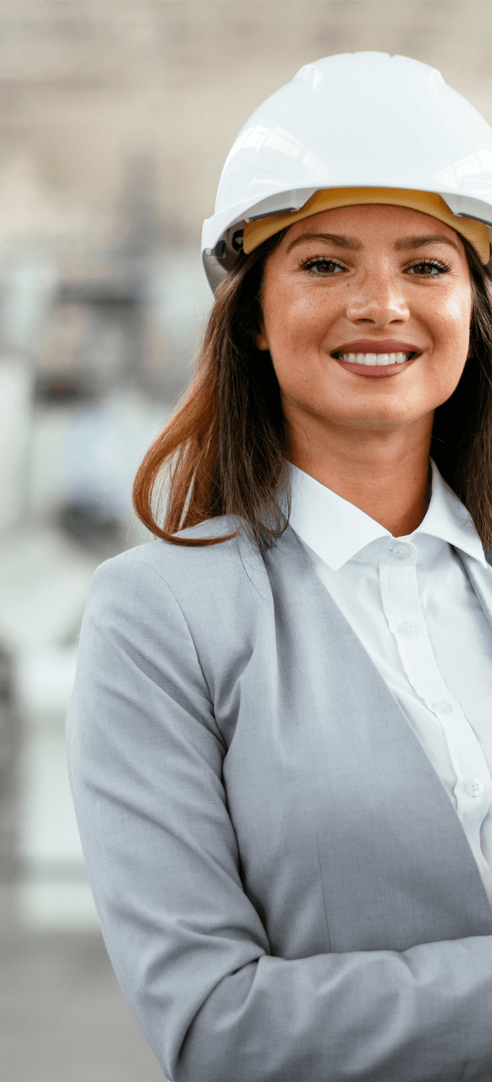 A woman wearing a white safety helmet and light grey suit. In the background, out of focus, is inside a factory.